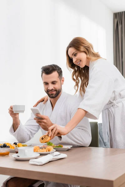 Donna sorridente in accappatoio prendere pasticceria vicino fidanzato utilizzando smartphone e tenendo tazza in camera d'albergo — Foto stock