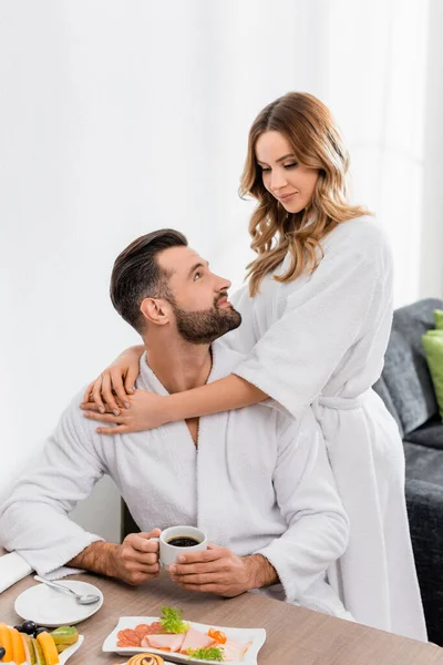 Mulher de roupão abraçando namorado com xícara de café perto do café da manhã em primeiro plano desfocado no hotel — Fotografia de Stock