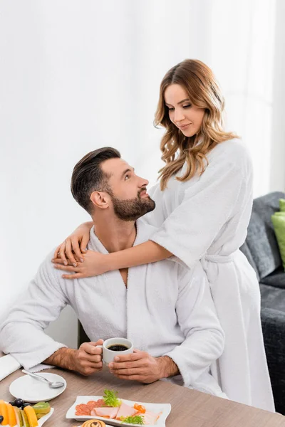 Frau im Bademantel umarmt Freund mit Tasse Kaffee beim Frühstück auf verschwommenem Vordergrund im Hotel — Stockfoto