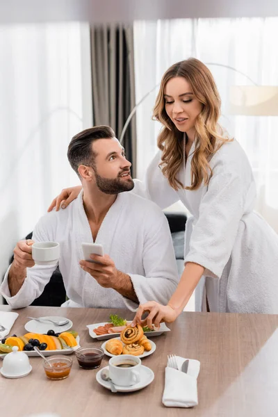 Woman taking pastry near boyfriend with cup of coffee and cellphone in hotel room — Stock Photo