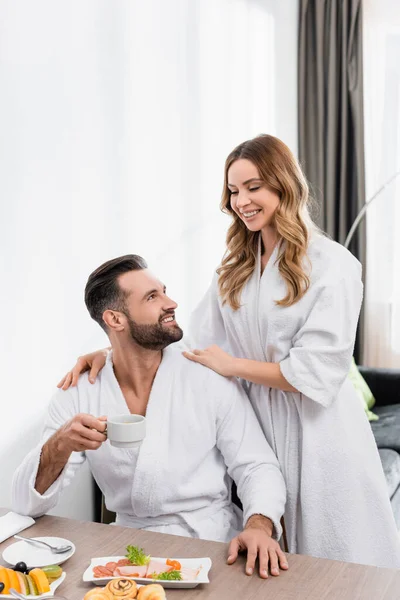 Smiling woman in white bathrobe embracing boyfriend with cup near tasty breakfast on table in hotel — Stock Photo