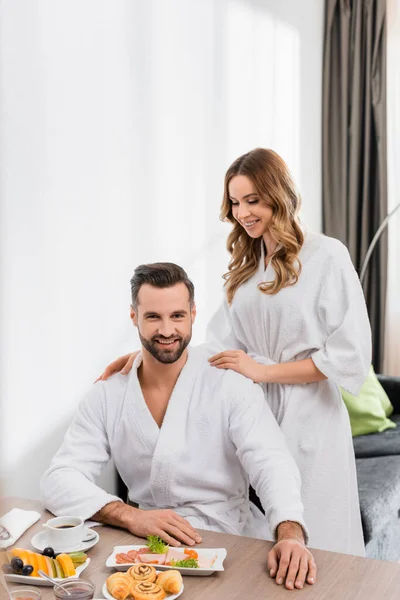 Cheerful woman in bathrobe hugging boyfriend near breakfast with coffee on table in hotel room — Stock Photo