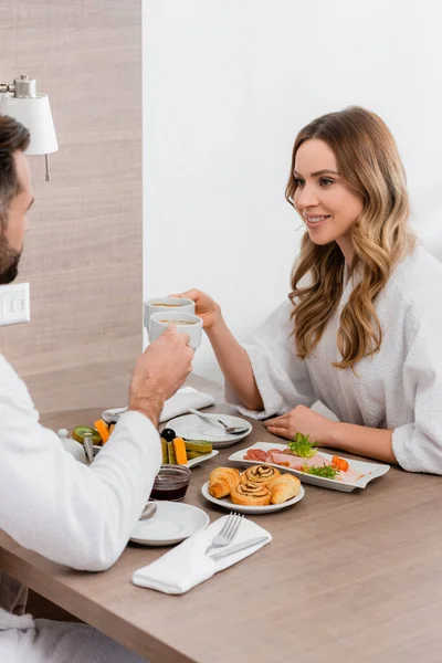 Smiling woman in bathrobe holding cup of coffee near breakfast and boyfriend on blurred foreground in hotel room — Stock Photo