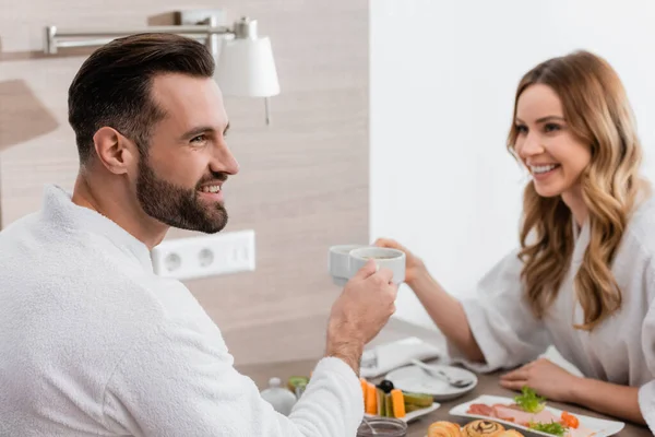 Smiling man in bathrobe holding cup near breakfast and girlfriend on blurred background in hotel — Stock Photo