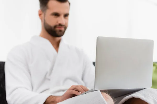 Laptop em voltas de homem de roupão de banho em fundo embaçado no quarto de hotel — Fotografia de Stock