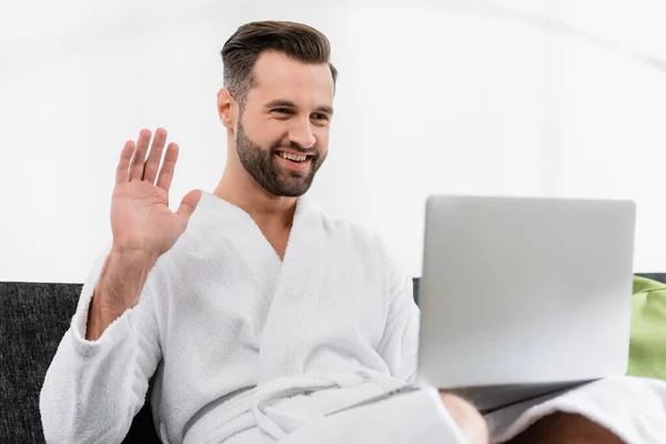 Homme joyeux en peignoir ayant appel vidéo sur ordinateur portable au premier plan flou dans la chambre d'hôtel — Photo de stock