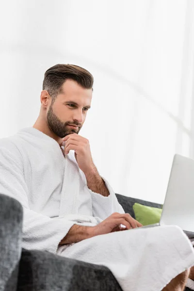 Libero professionista in accappatoio con computer portatile sul divano in primo piano sfocato in hotel — Foto stock