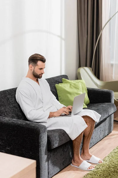 Телеработник в тапочках и халате с помощью ноутбука на диване в отеле — Stock Photo