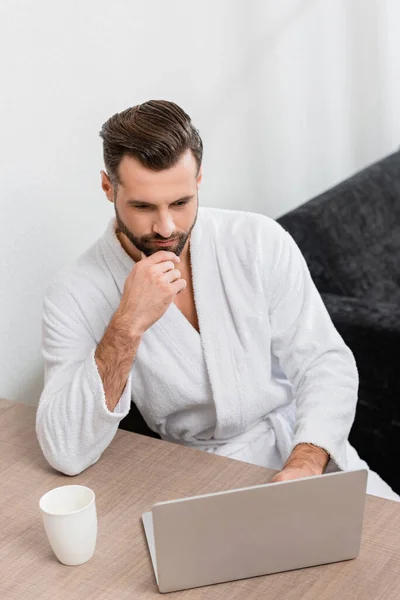 Freelancer in bathrobe looking at laptop near cup in hotel — Stock Photo