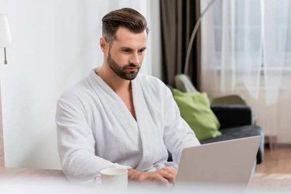 Hombre barbudo en albornoz usando el ordenador portátil cerca de la taza en primer plano borroso en la habitación del hotel - foto de stock