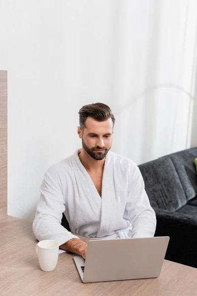 Homme en peignoir blanc avec ordinateur portable près de la tasse sur la table dans la chambre d'hôtel — Photo de stock