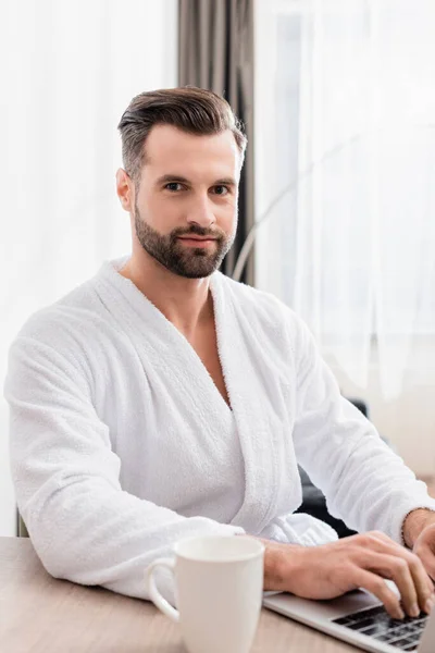 Man in bathrobe looking at camera while using laptop near cup on burred foreground in hotel — Stock Photo