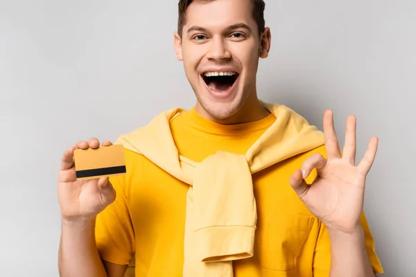 Homem alegre com cartão de crédito mostrando gesto ok em fundo cinza — Fotografia de Stock