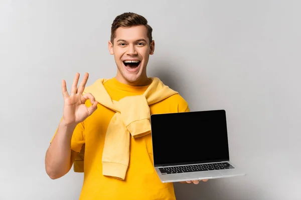 Hombre positivo sosteniendo el ordenador portátil con pantalla en blanco y mostrando gesto aceptable sobre fondo gris - foto de stock