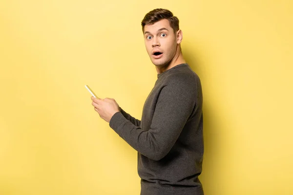 Amazed man looking at camera while using smartphone on yellow background — Stock Photo