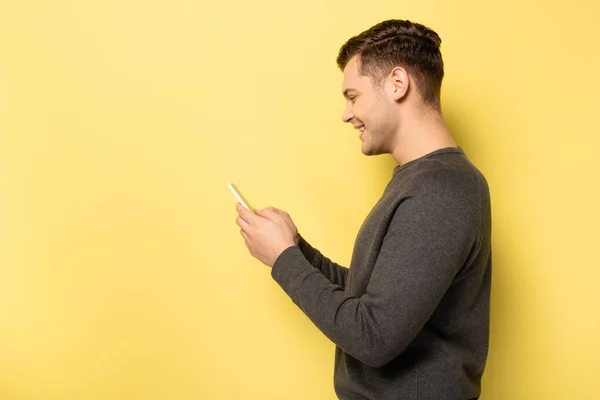 Side view of smiling man in grey pullover using cellphone on yellow background — Stock Photo