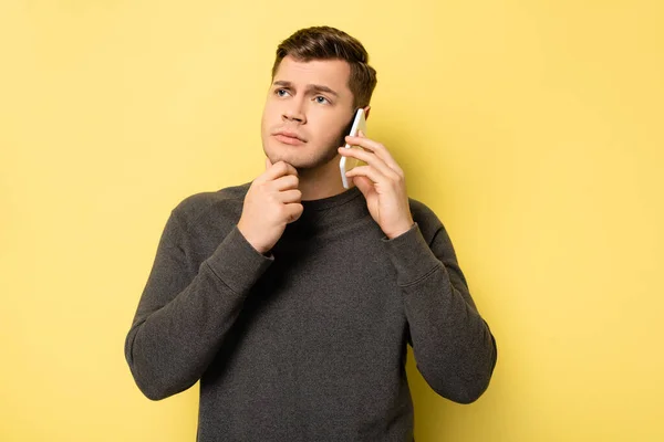 Hombre pensativo con la mano cerca de la barbilla hablando en el teléfono inteligente sobre fondo amarillo - foto de stock