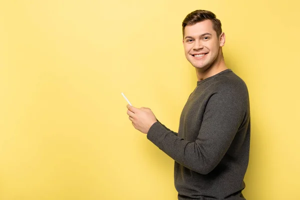 Homme souriant utilisant un téléphone mobile et regardant la caméra sur fond jaune — Photo de stock