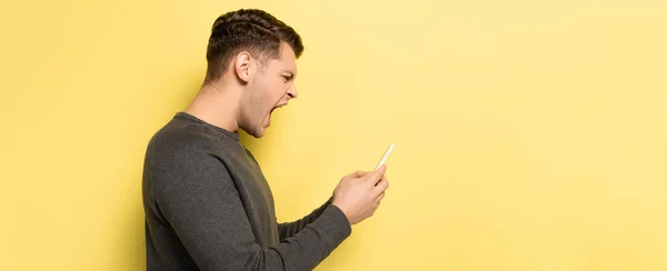 Side view of angry man screaming while using smartphone on yellow background, banner — Stock Photo