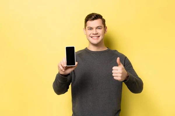 Homem mostrando smartphone e polegar no fundo amarelo — Fotografia de Stock