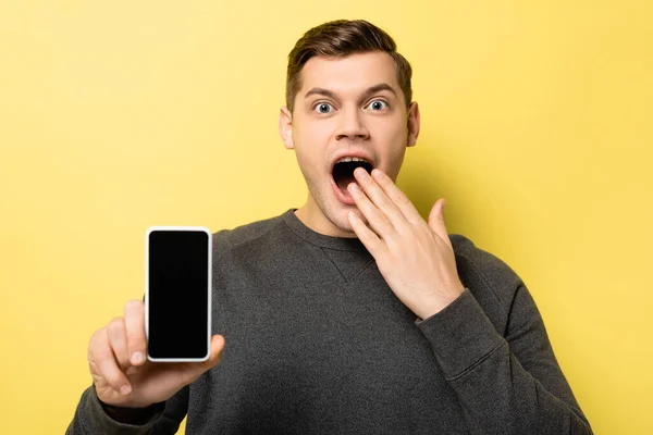 Uomo stupito che mostra smartphone con schermo bianco su sfondo giallo — Foto stock