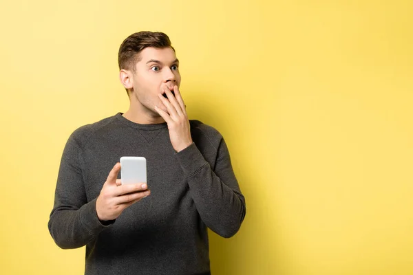 Aufgeregter Mann mit Hand in Mundnähe und Handy auf gelbem Hintergrund — Stockfoto