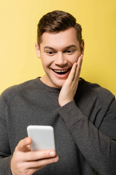Happy man with hand near cheek holding smartphone on blurred foreground on yellow background — Stock Photo