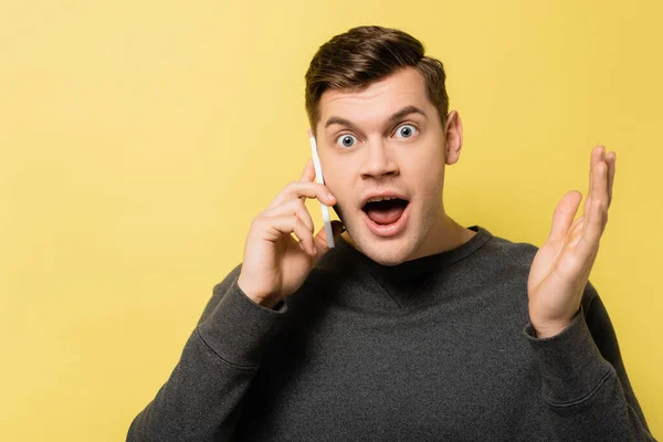 Excited man looking at camera while talking on smartphone on yellow background — Stock Photo