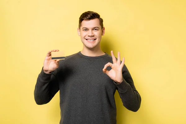 Fröhlicher Mann mit Kreditkarte zeigt ok Geste auf gelbem Hintergrund — Stockfoto