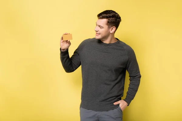 Homme souriant avec la main dans la poche regardant la carte de crédit sur fond jaune — Photo de stock