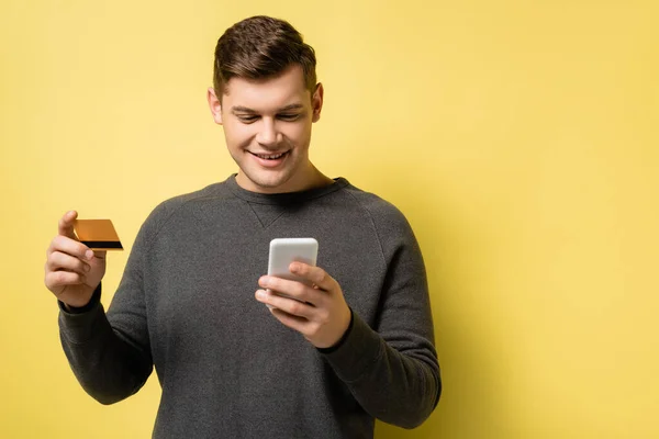 Hombre sonriente usando teléfono inteligente y tarjeta de crédito sobre fondo amarillo - foto de stock