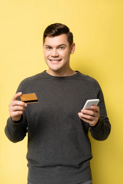 Lächelnder Mann mit Kreditkarte und Smartphone vor der Kamera auf gelbem Hintergrund — Stockfoto