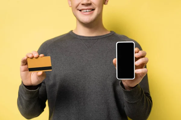 Cropped view of smartphone with blank screen and credit card in hands of smiling man blurred on yellow background — Stock Photo