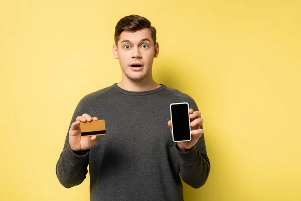 Hombre sorprendido sosteniendo tarjeta de crédito y teléfono inteligente sobre fondo amarillo - foto de stock