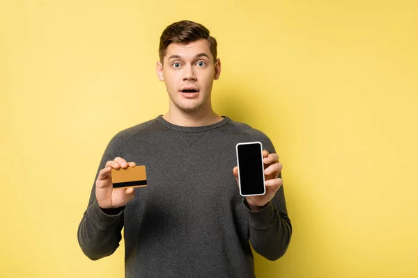 Homem surpreendente segurando cartão de crédito e smartphone no fundo amarelo — Fotografia de Stock