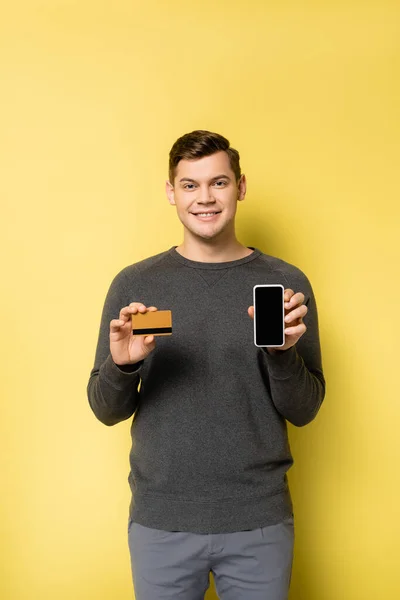Uomo sorridente che mostra carta di credito e smartphone su sfondo giallo — Foto stock