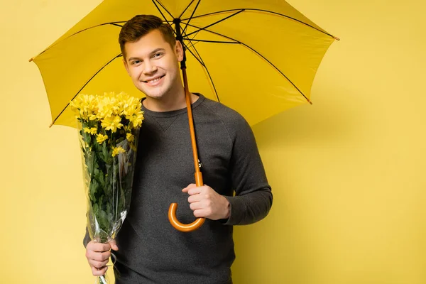 Homme souriant avec des fleurs et un parapluie regardant la caméra sur fond jaune — Photo de stock