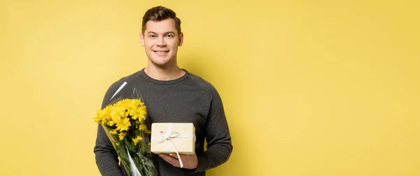 Junger Mann lächelt, während er Geschenke und Blumen auf gelbem Hintergrund hält, Banner — Stockfoto