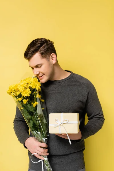 Mann im grauen Pullover lächelt, während er Geschenkschachtel und Blumenstrauß auf gelbem Hintergrund hält — Stockfoto
