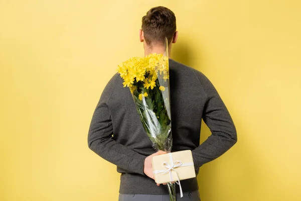 Back view of man hiding flowers and gift on yellow background — Stock Photo