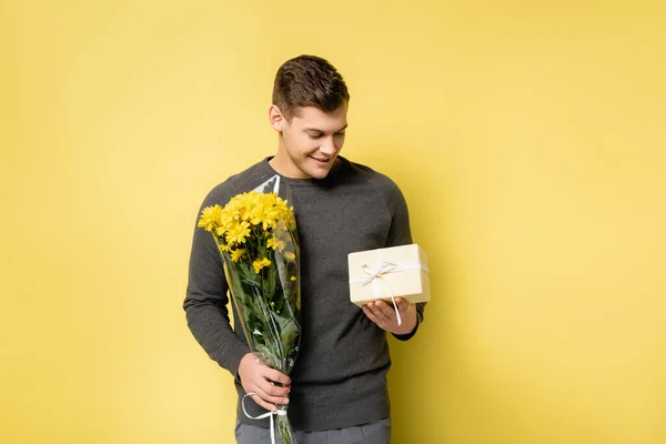 Lächelnder Mann betrachtet Geschenk und hält Blumen auf gelbem Hintergrund — Stockfoto