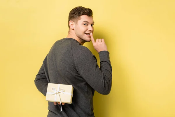 Back view of man showing shh gesture and hiding present on yellow background — Stock Photo