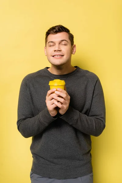 Happy man with closed eyes holding coffee to go on yellow background — Stock Photo