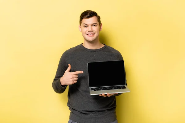 Homme souriant pointant vers un ordinateur portable avec écran blanc sur fond jaune — Photo de stock