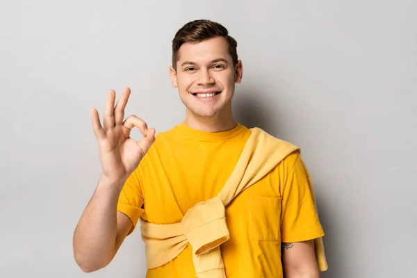 Smiling man in yellow clothes showing ok gesture on grey background — Stock Photo