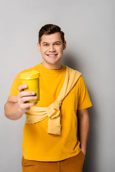 Hombre sonriente con la mano en el bolsillo sosteniendo café para ir en primer plano borroso sobre fondo gris - foto de stock