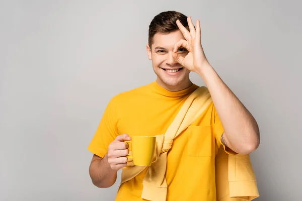 Hombre sonriente con taza mostrando gesto aceptable sobre fondo gris - foto de stock