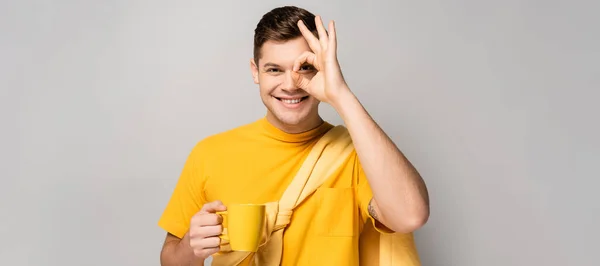 Uomo allegro mostrando simbolo ok e tenendo la tazza su sfondo grigio, banner — Foto stock