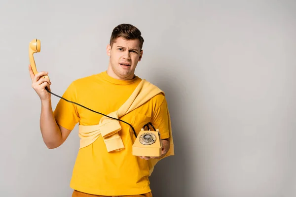 Homme gêné tenant un téléphone rétro jaune sur fond gris — Photo de stock