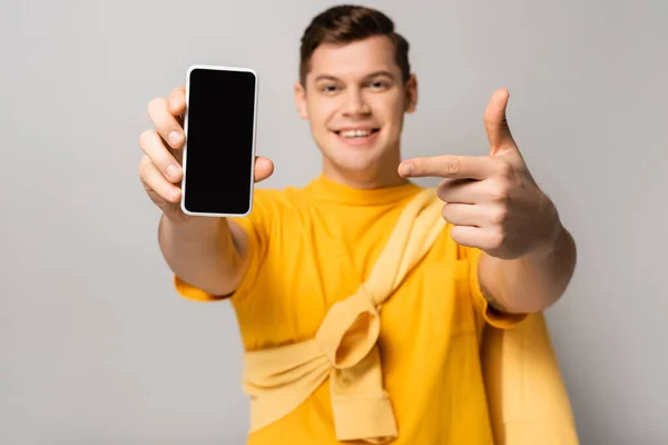 Smartphone avec écran blanc dans la main de l'homme souriant pointant avec le doigt flou sur fond gris — Photo de stock